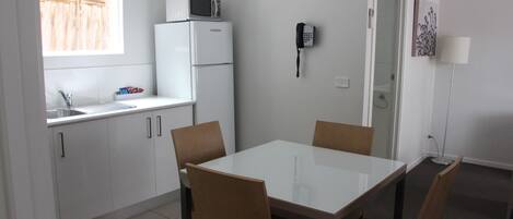 Cocina compacta con microondas, refrigerador y teléfono en la pared. Hay una mesa de comedor con sillas y una ventana con persianas.