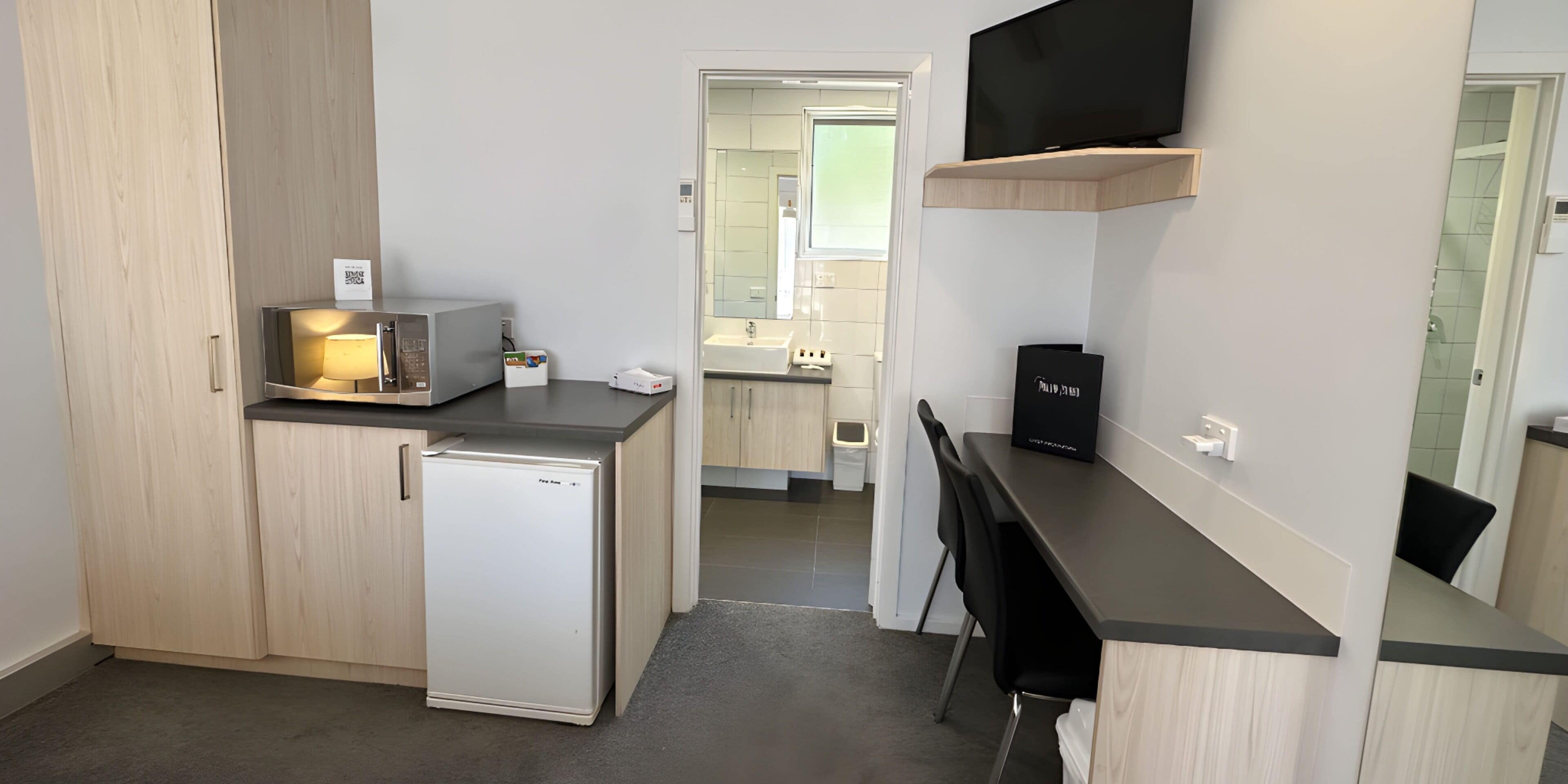 Una cocina compacta con microondas, refrigerador y un lavamanos pequeño. Hay un televisor montado en la pared y una silla en la barra.