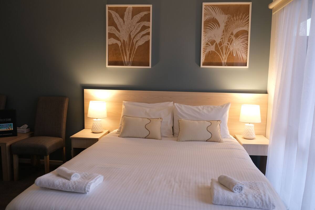 A neatly made bed with white linens and pillows, two wall-mounted lamps, and a framed plant artwork above the bed.