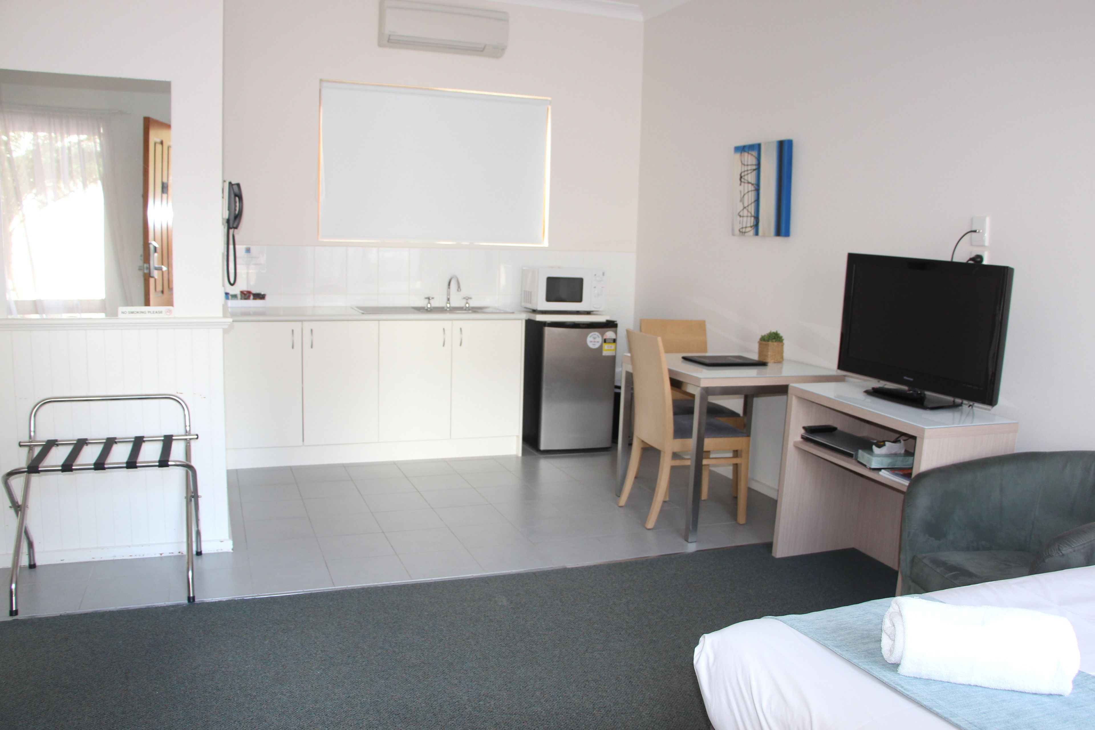 A modern hotel room with a bed, a desk with a computer and TV, a small kitchenette, and a bathroom.