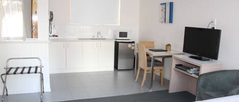 A modern hotel room with a bed, a desk with a computer and TV, a small kitchenette, and a bathroom.