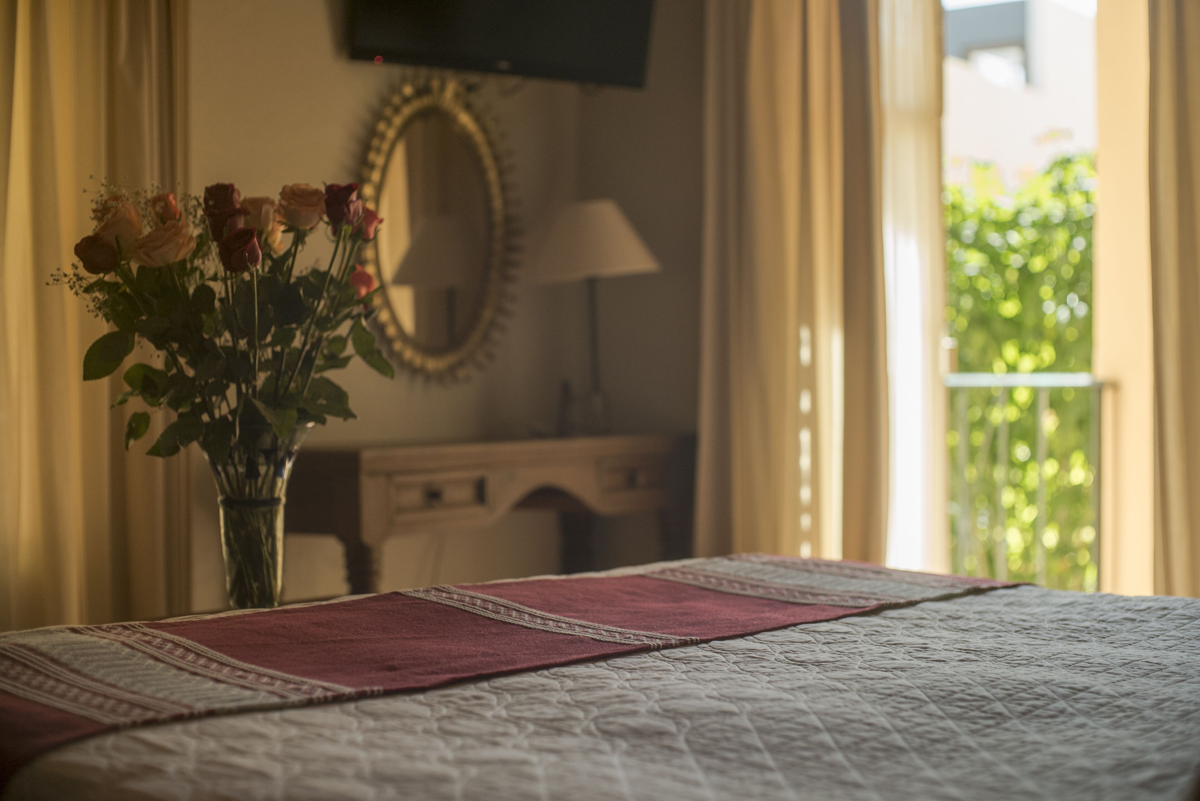A bed with a quilt and a red runner, a vase with flowers, a mirror, and a lamp.