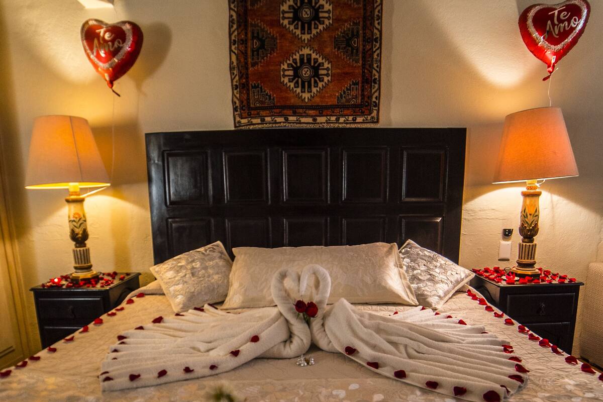 A neatly made bed with towels folded in a heart shape, a vase with a single red rose, and heart-shaped balloons on the nightstands.