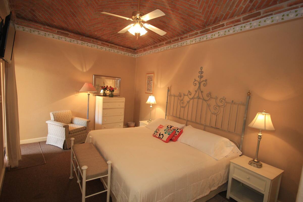 A bedroom with a large bed, a nightstand with a lamp, a chair, a dresser, and a ceiling fan.