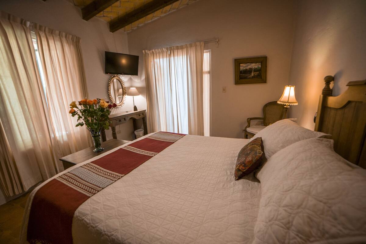 A hotel room with a large bed, a desk with a chair, a television mounted on the wall, and a window with curtains.