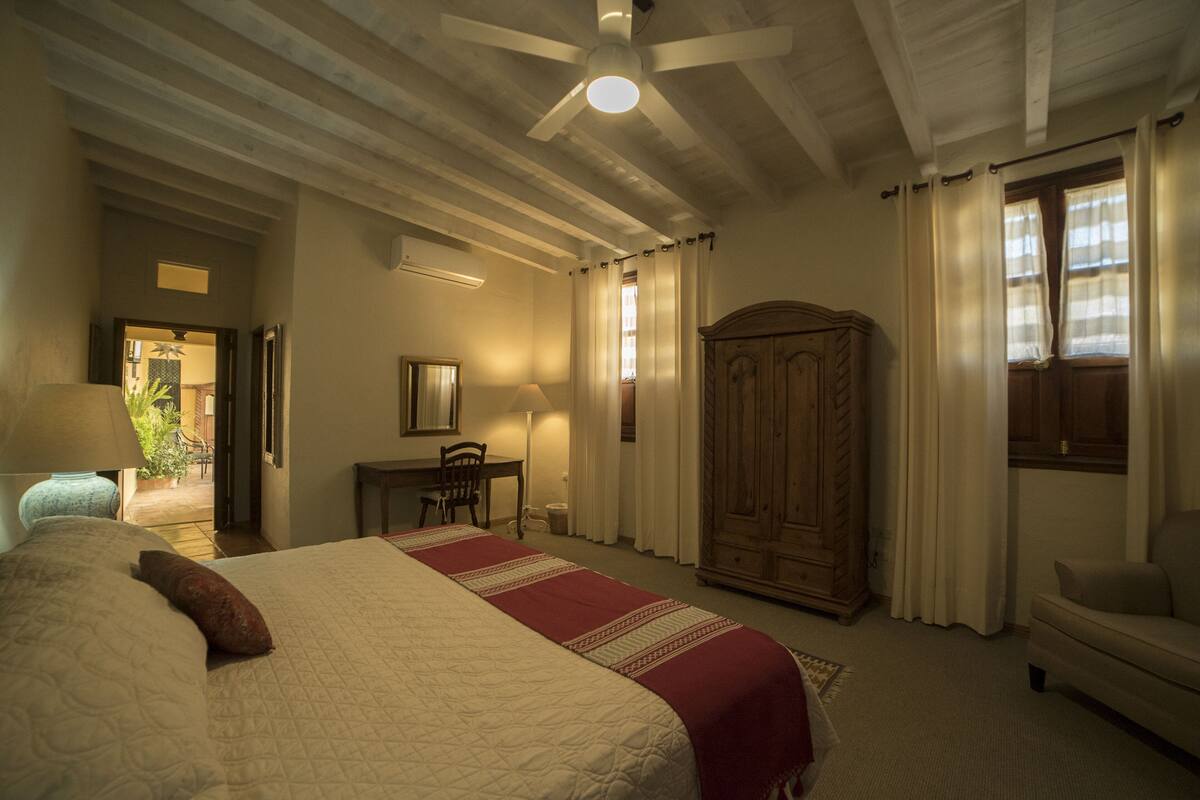 A bedroom with a large bed, a wooden wardrobe, a desk with a chair, and a ceiling fan.