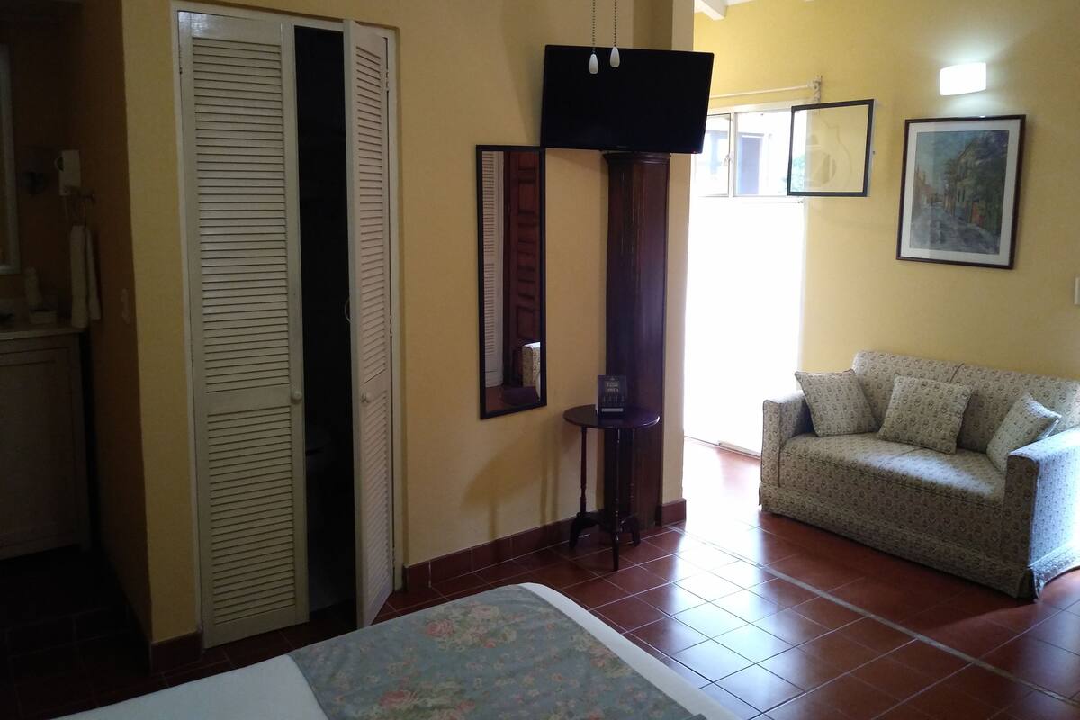 A room with a bed, a sofa, a TV mounted on the wall, and a door with louvered shutters.