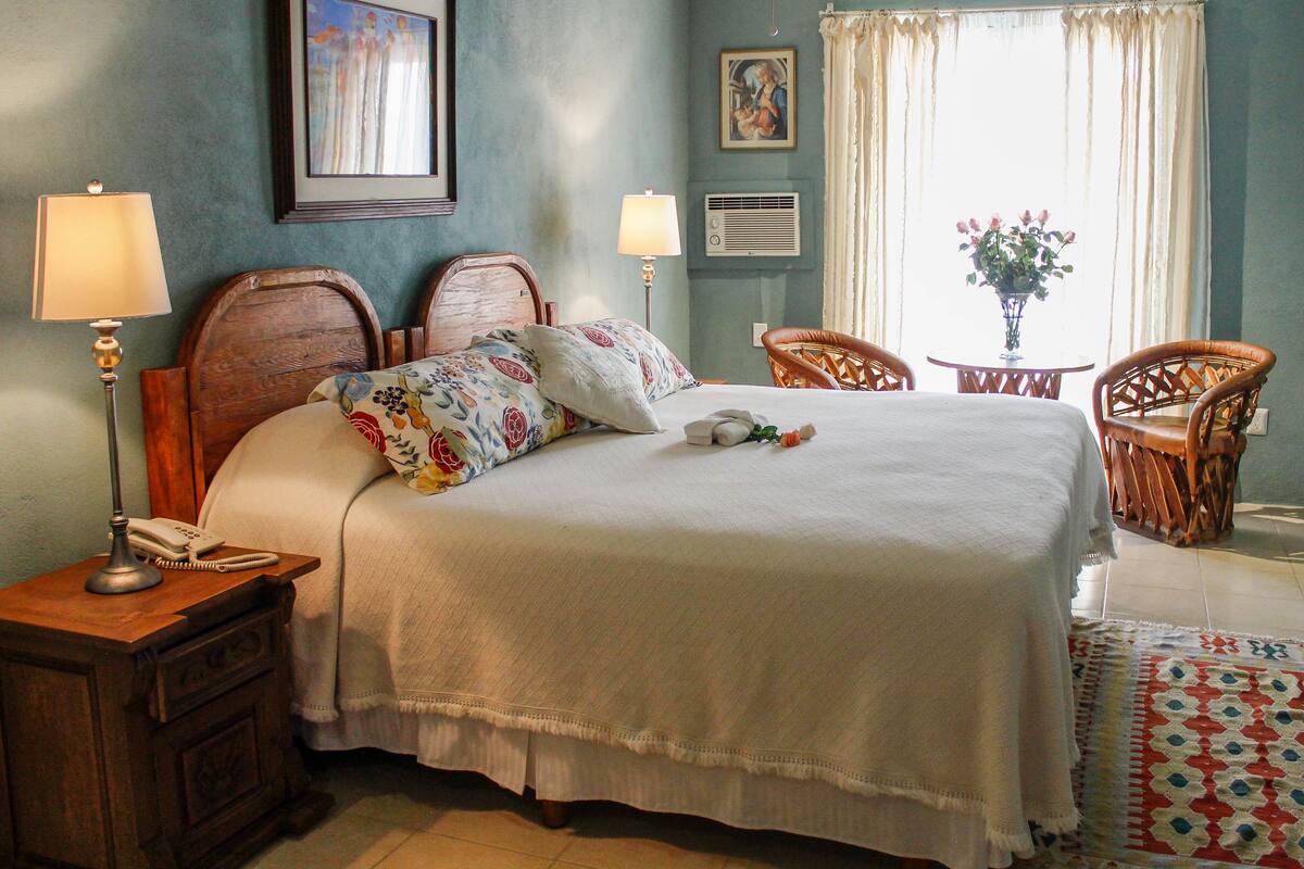 A neatly made bed with floral pillows, a bedside table with a lamp and phone, and a chair by the window.