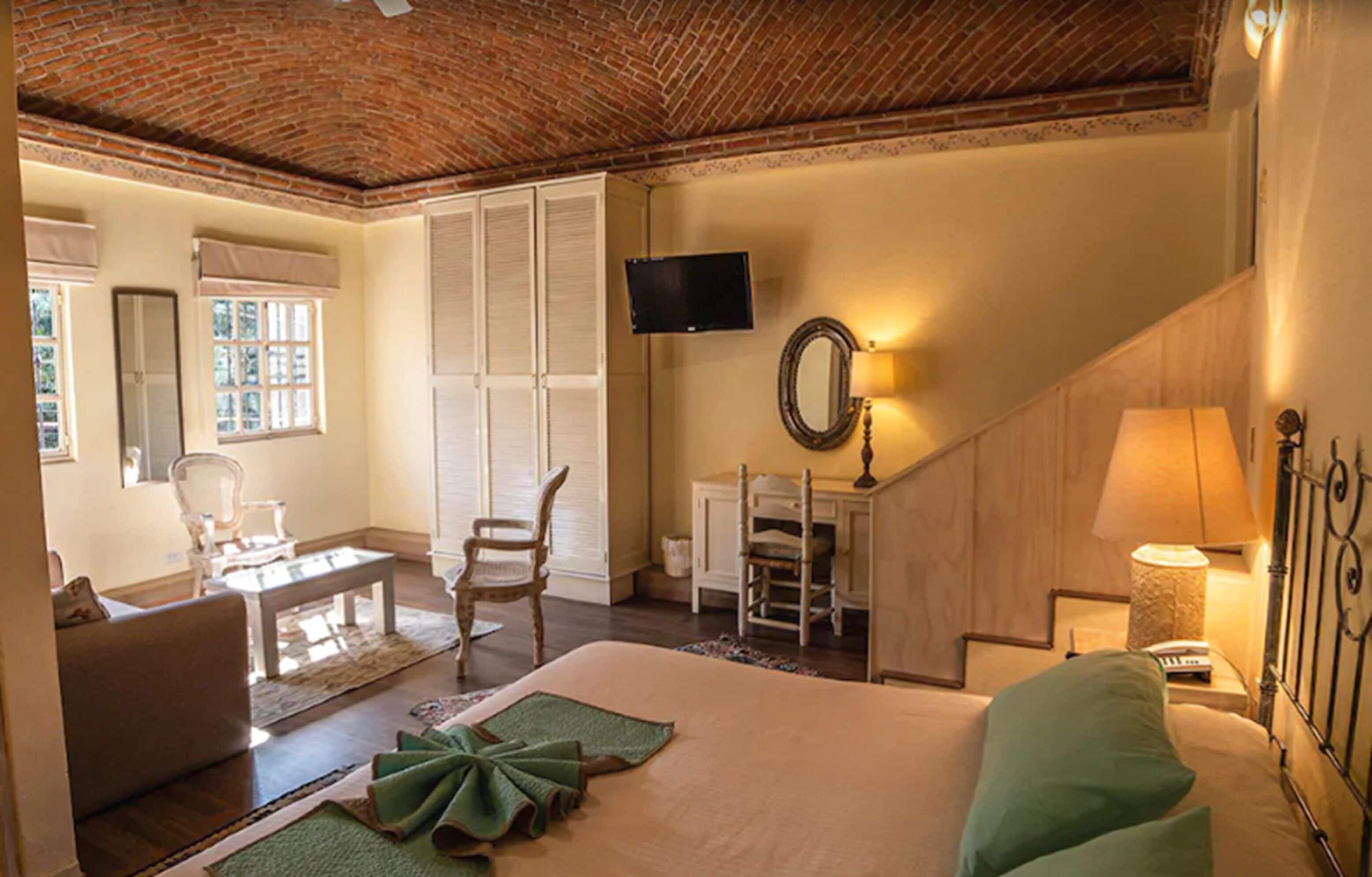 Habitación de hotel con techo de ladrillo, cama con almohadas verdes, escritorio con lámpara, una silla y televisor montado en la pared.