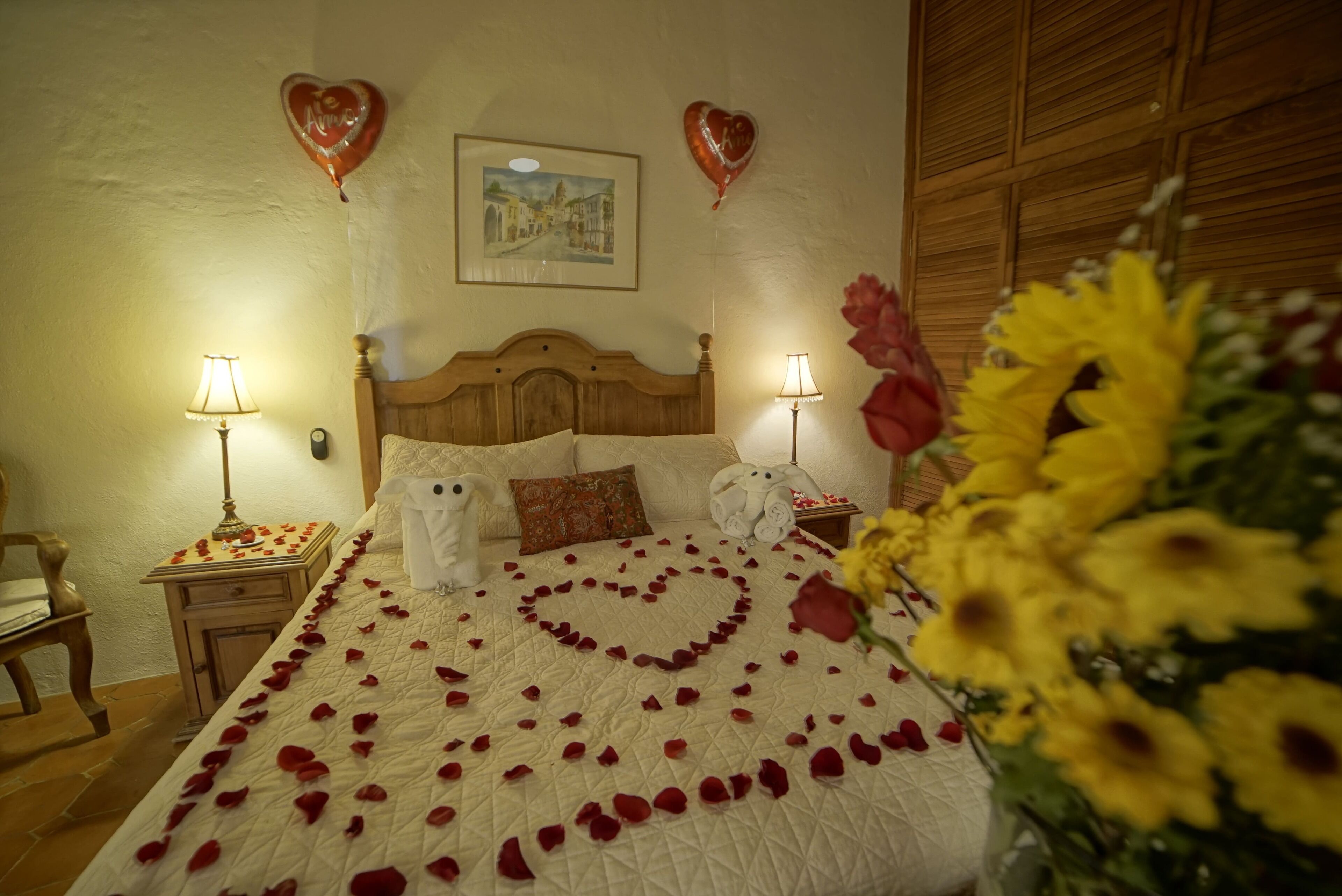 A hotel room with a bed adorned with rose petals forming a heart shape, two heart-shaped balloons, and a bouquet of sunflowers.