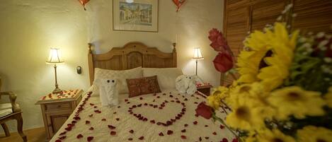 A hotel room with a bed adorned with rose petals forming a heart shape, two heart-shaped balloons, and a bouquet of sunflowers.
