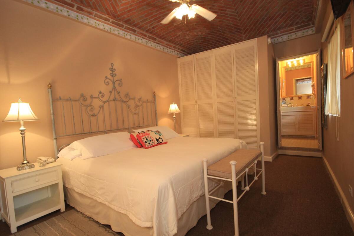 A bedroom with a bed, nightstands, a ceiling fan, and a bathroom with a mirror and sink.