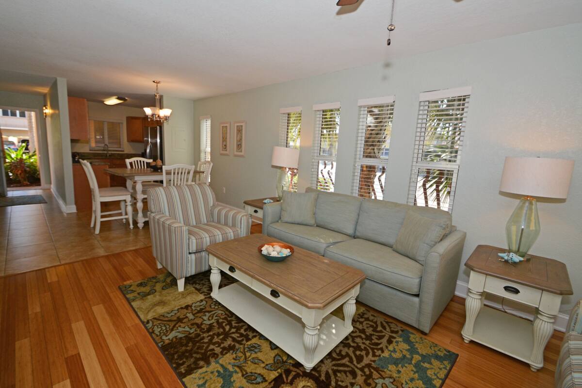A living room with a sofa, chairs, a coffee table, and a lamp. There is a kitchen in the background.