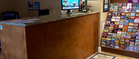 Un área de recepción con computadora, escritorio, silla, decoración en la pared, lámpara y una exhibición de libros.