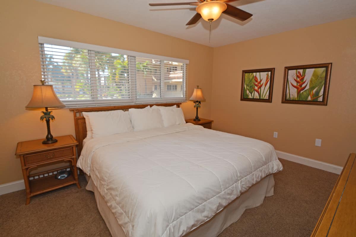 A bedroom with a large bed, two bedside tables with lamps, a ceiling fan, and two framed pictures on the wall.