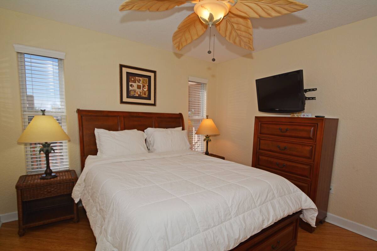 A bedroom with a wooden bed, two bedside tables with lamps, a ceiling fan, a TV on a dresser, and a framed picture on the wall.