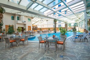 An indoor swimming pool area with multiple lounge chairs and tables, surrounded by potted plants and a glass ceiling. - Bristol Hotel (Amman)