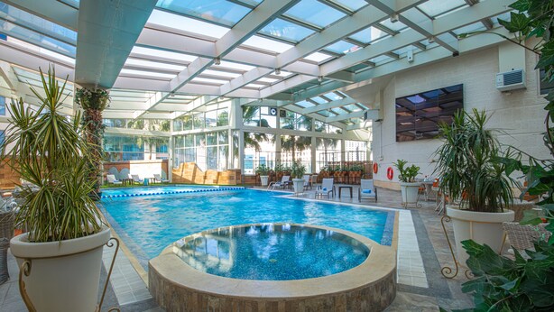 An indoor swimming pool with a glass ceiling, surrounded by potted plants and lounge chairs. - Bristol Hotel (Amman)