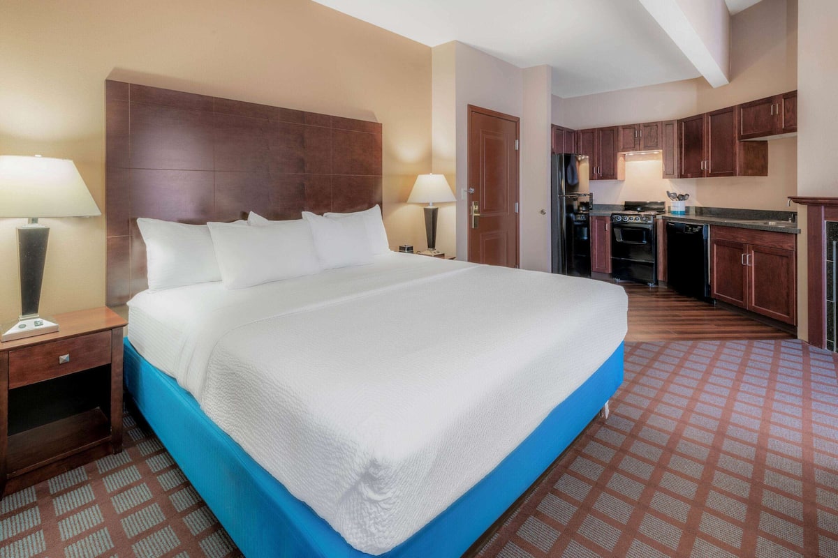 A hotel room with a large bed, a nightstand, a desk, a lamp, a wardrobe, and a kitchenette.