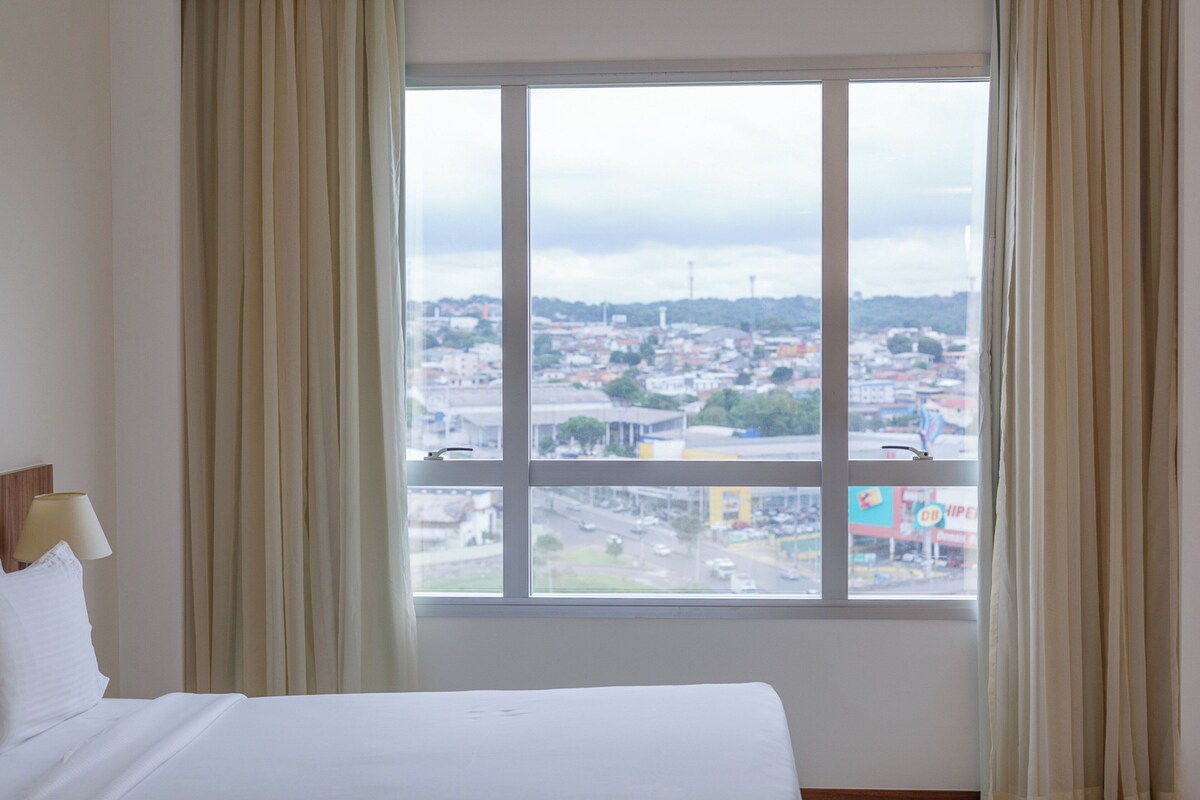 Habitación de hotel con cama, ventana con cortinas y vistas a la ciudad.