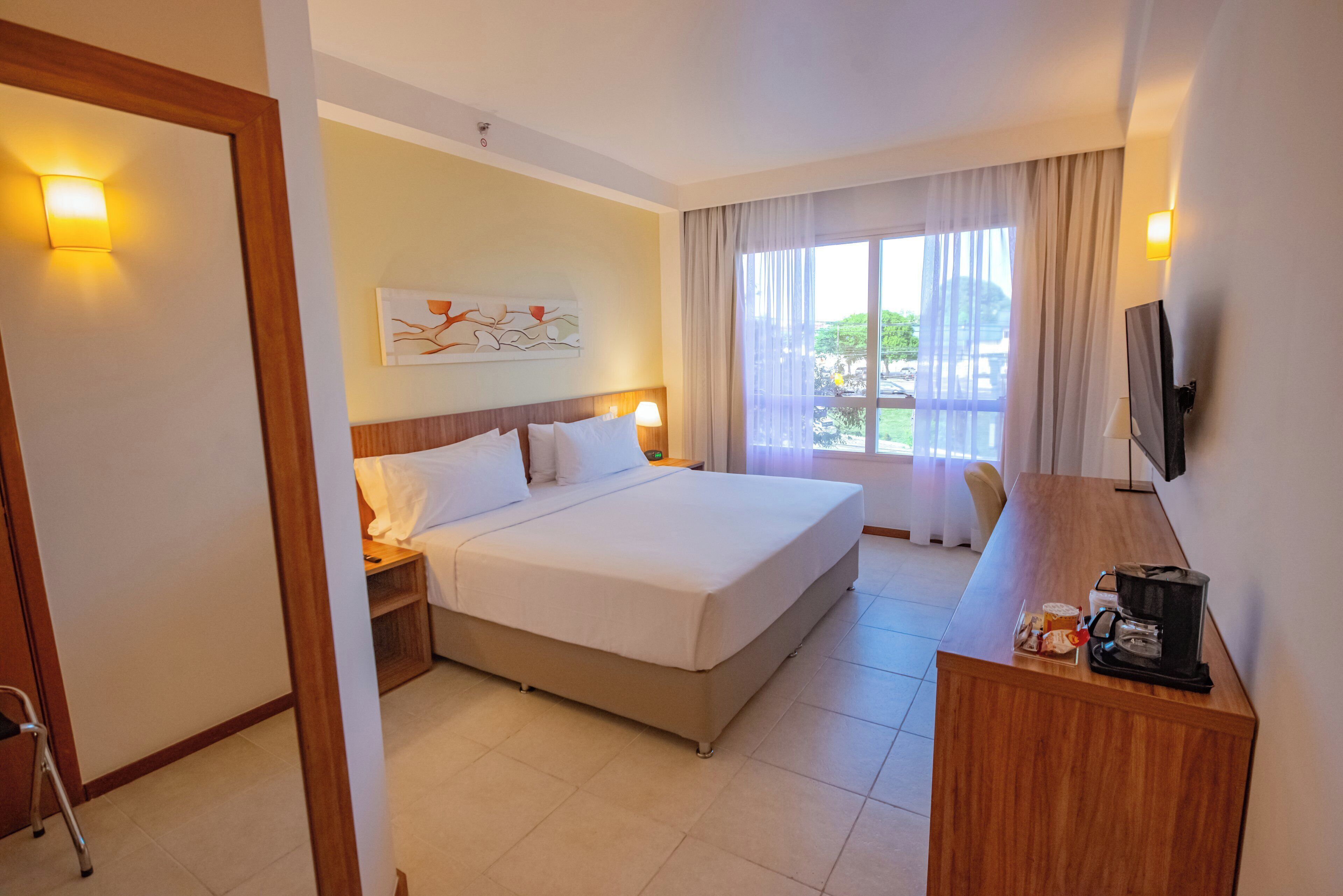 Quarto de hotel com uma cama grande, mesas de cabeceira, escrivaninha com cafeteira, televisão e janela com cortinas.