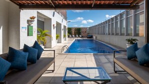 Un área junto a la piscina con suelo de baldosas, mobiliario de relax con cojines azules, una mesa de cristal y vistas despejadas a la piscina.