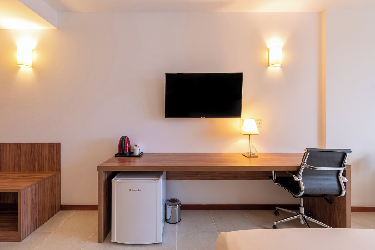 Une chambre d’hôtel avec un bureau en bois, un téléviseur fixé au mur, un petit réfrigérateur, une poubelle, une chaise et une table de chevet.