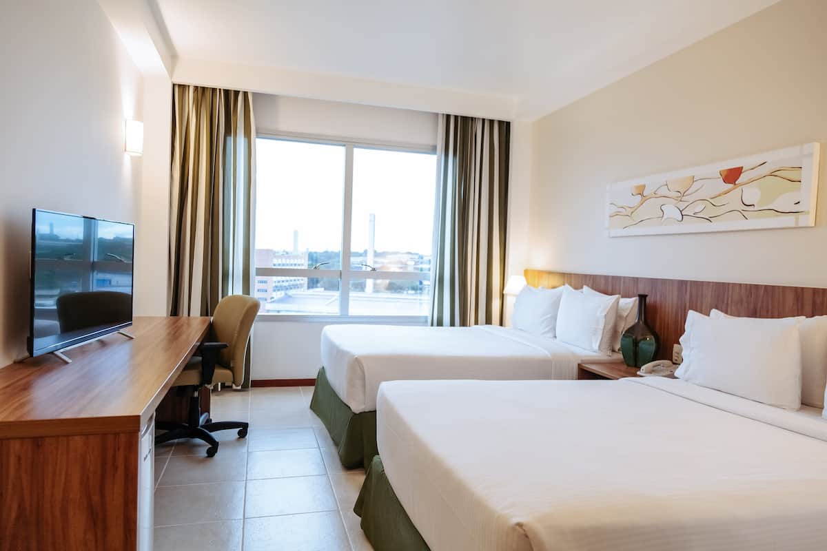 A hotel room with two beds, a desk with a TV, a chair, and a window with curtains.
