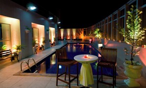 Un área de piscina nocturna con piscina rectangular, mobiliario para sentarse y plantas en macetas.