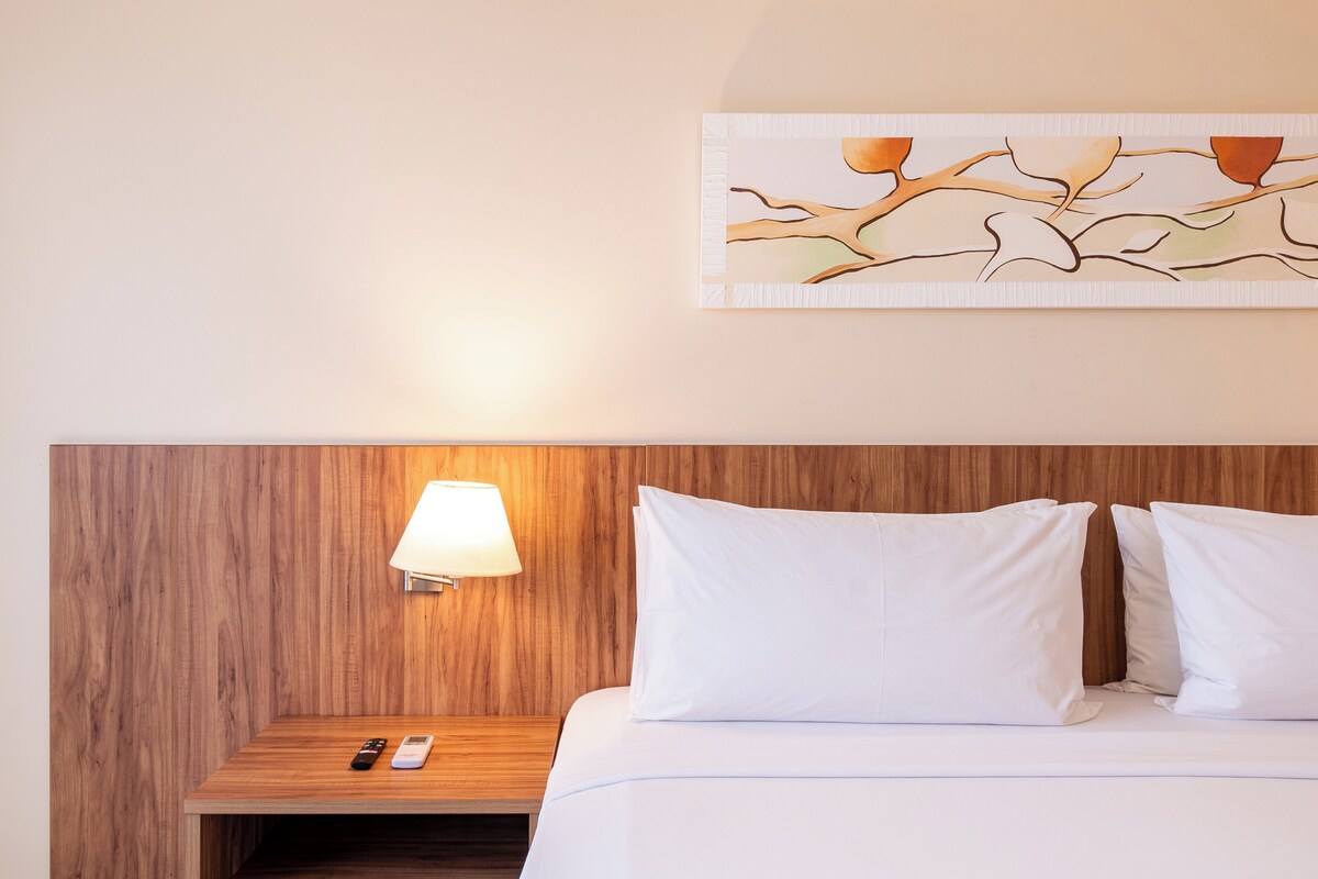 Une chambre d’hôtel avec une tête de lit en bois, une lampe de chevet, une table de chevet avec une télécommande et un lit blanc avec deux oreillers.