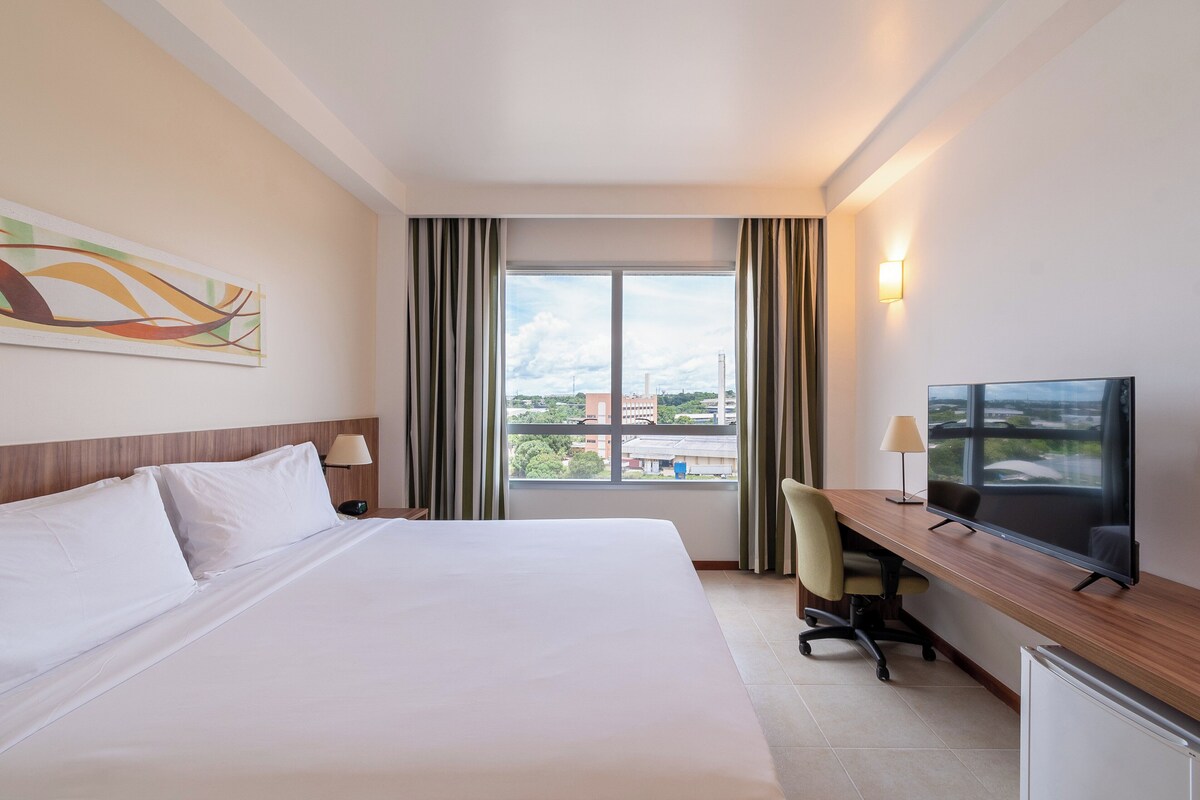 Habitación de hotel con cama, escritorio con televisor, una silla y ventana con cortinas.