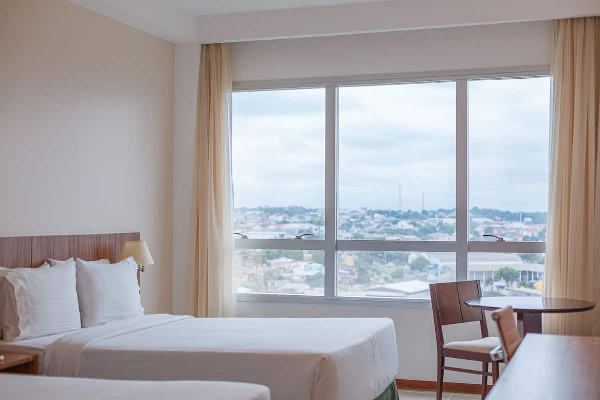 A hotel room with a bed, a chair, a table, and a large window offering a city view.