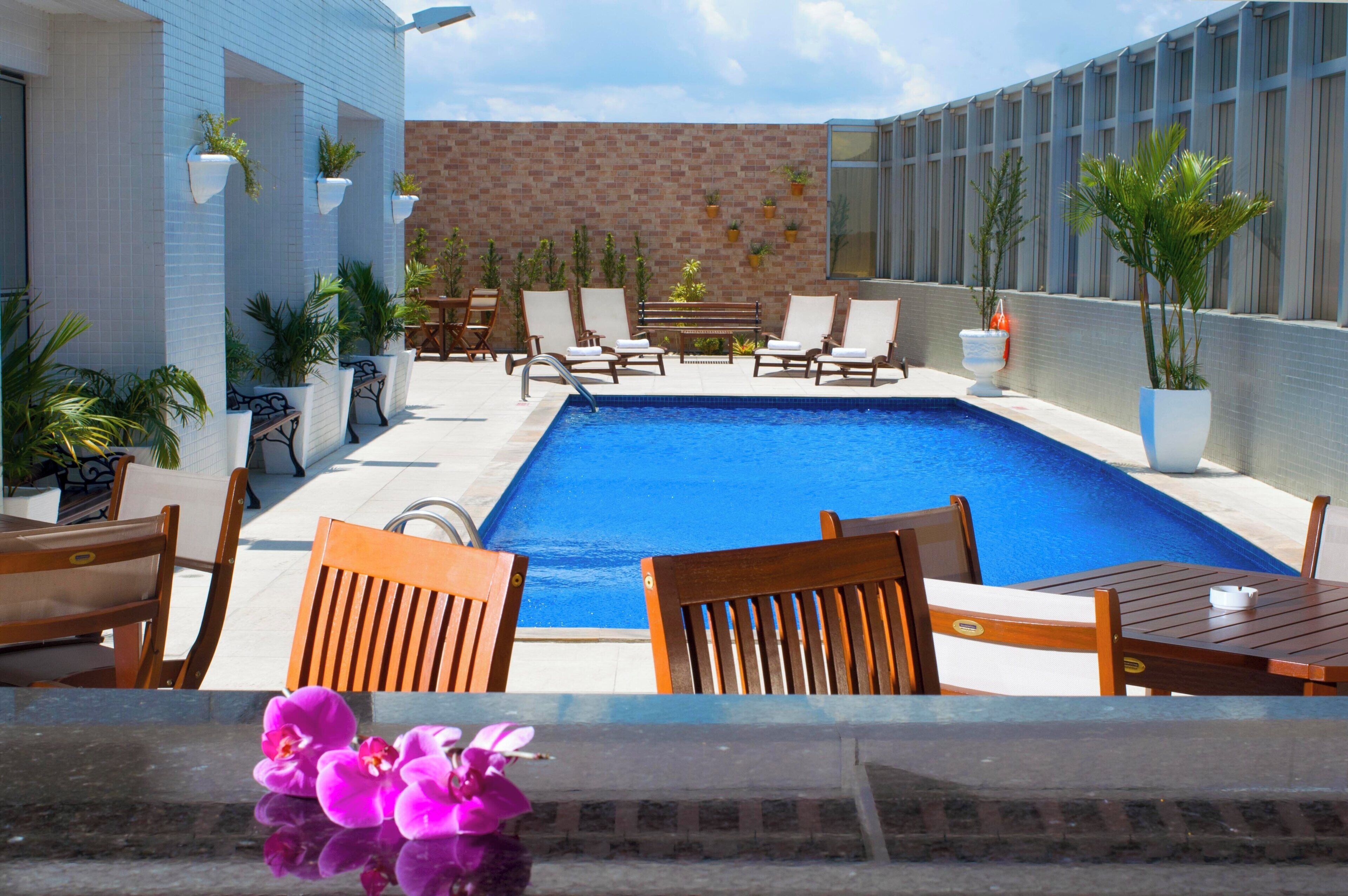 A swimming pool with lounge chairs and tables, surrounded by potted plants and a brick wall.