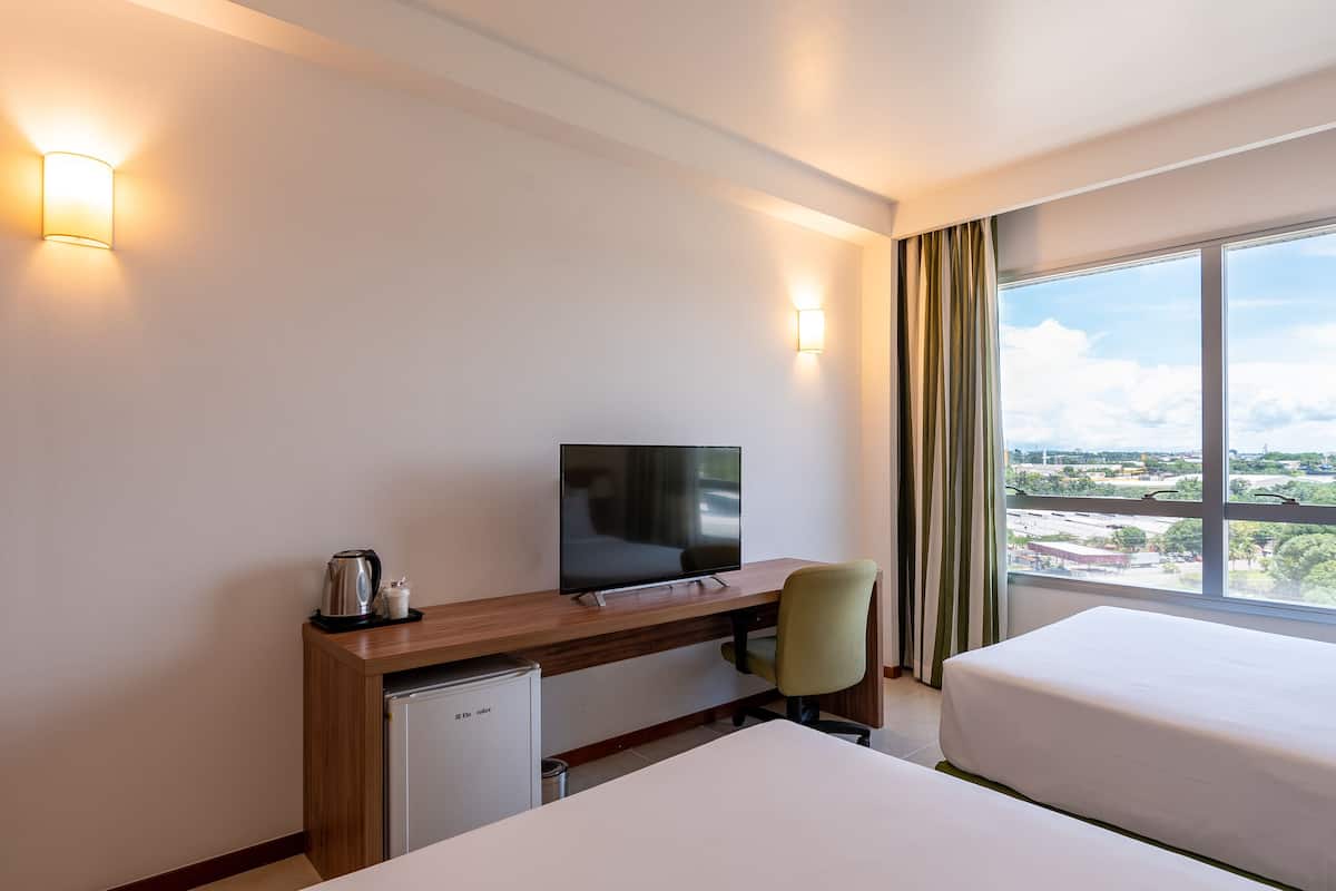 A hotel room with a bed, a desk with a television, a small refrigerator, and a view of the city through large windows.