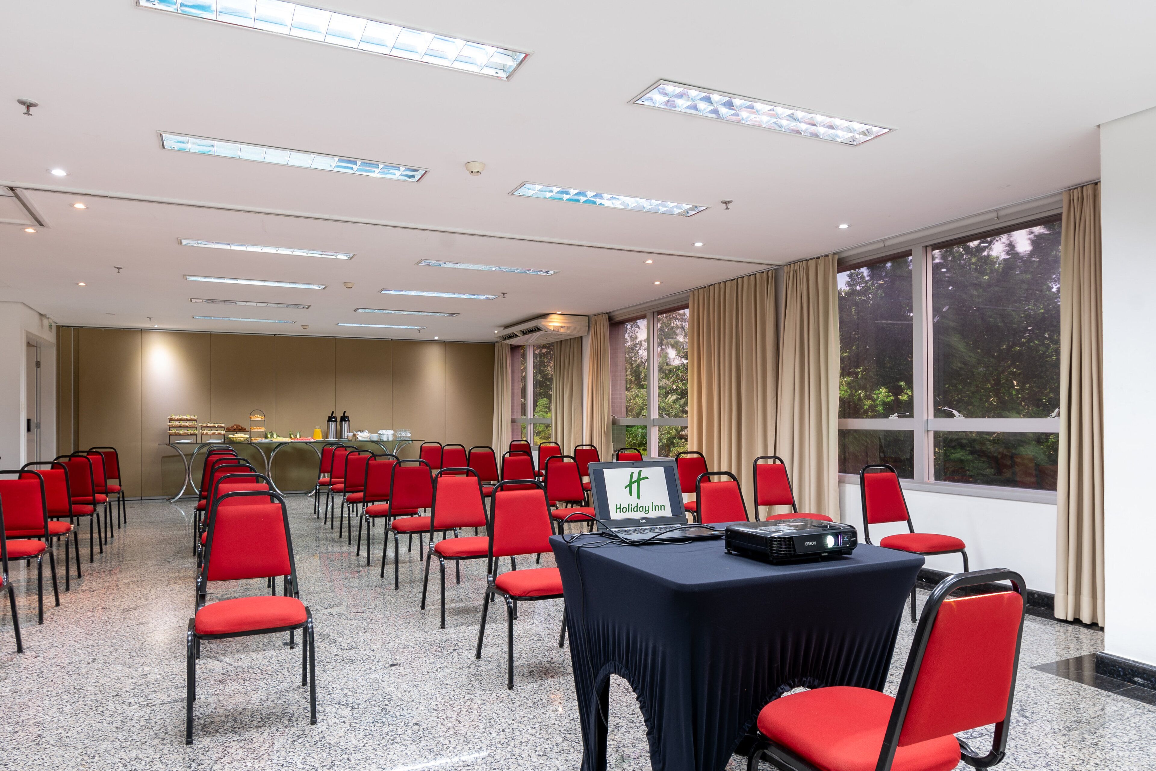 A conference room with red chairs, a table with a laptop, and a large window with curtains.