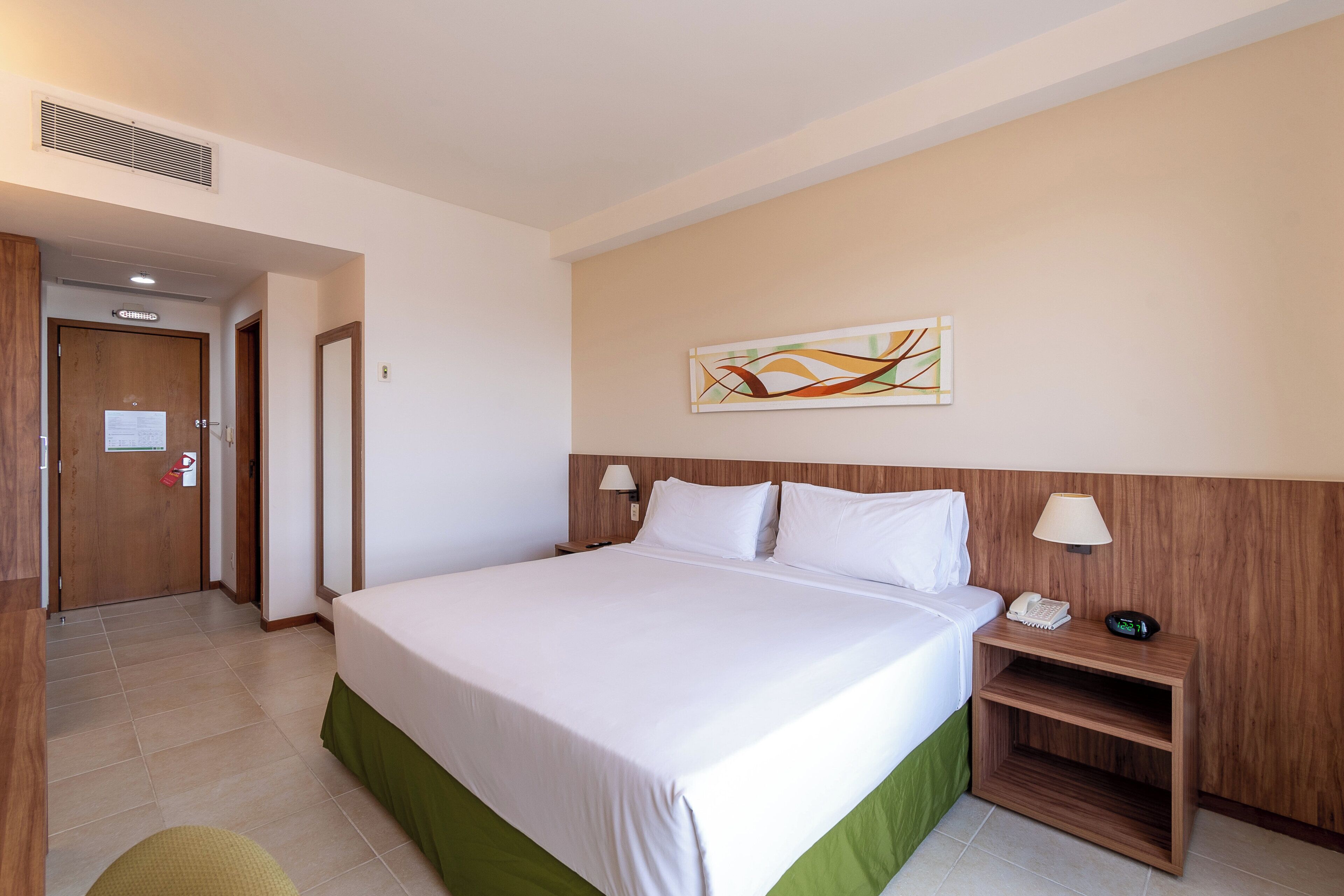 Habitación de hotel con cama, mesita de noche, lámpara de pared, un cuadro y una puerta que da a otra habitación.