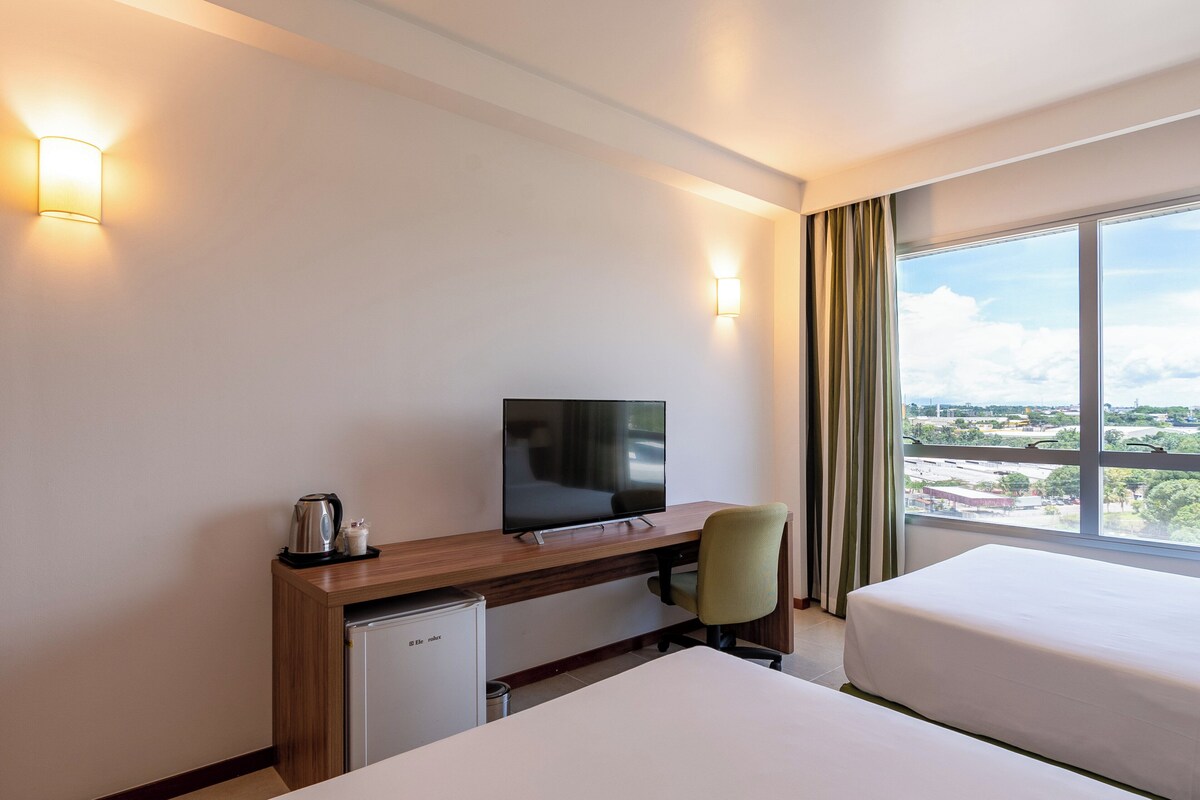 Une chambre d’hôtel comprenant un lit, un bureau avec une télévision, un petit réfrigérateur et une vue sur la ville grâce à de grandes fenêtres.