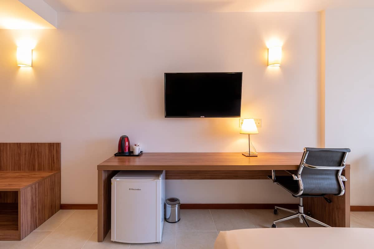 A hotel room with a wooden desk, a television mounted on the wall, a small refrigerator, a trash bin, a chair, and a bedside table.