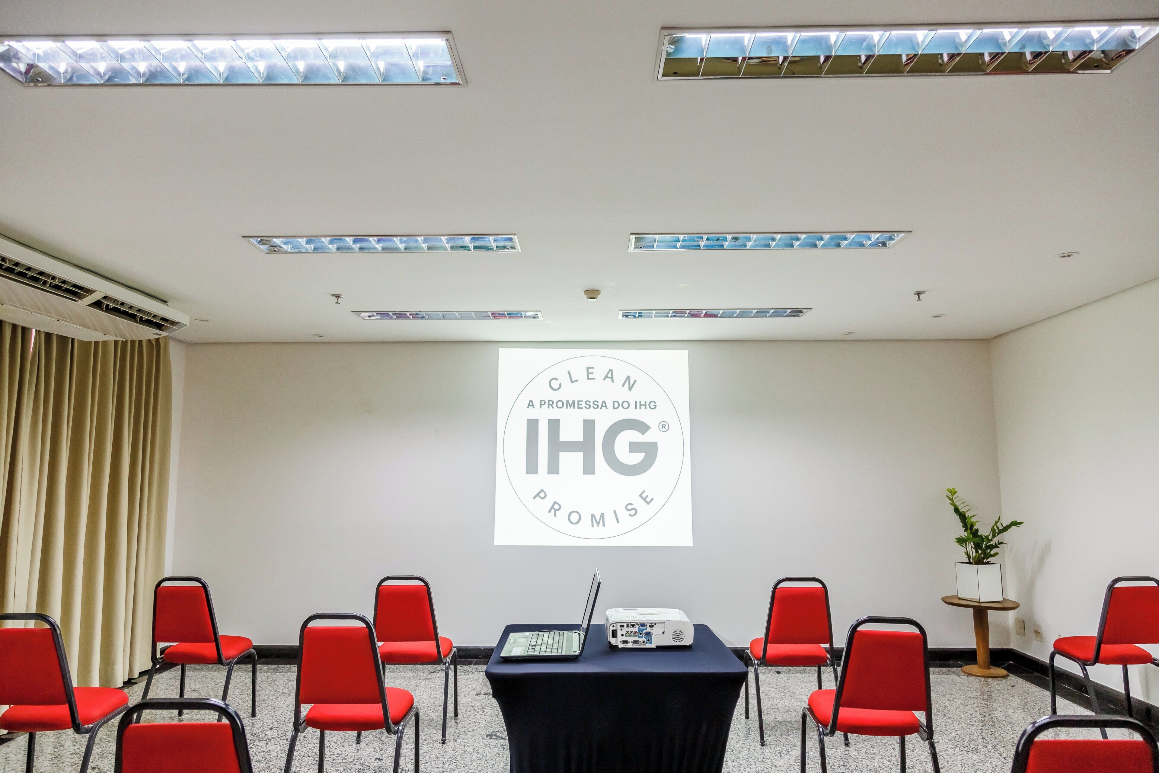 Un salón de conferencias con proyector, sillas rojas, una mesa y un cartel en la pared.