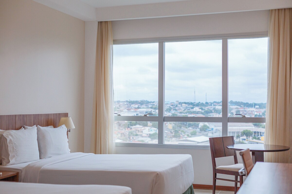 Une chambre d’hôtel équipée d’un lit, d’une chaise, d’une table et d’une grande fenêtre donnant sur la ville.
