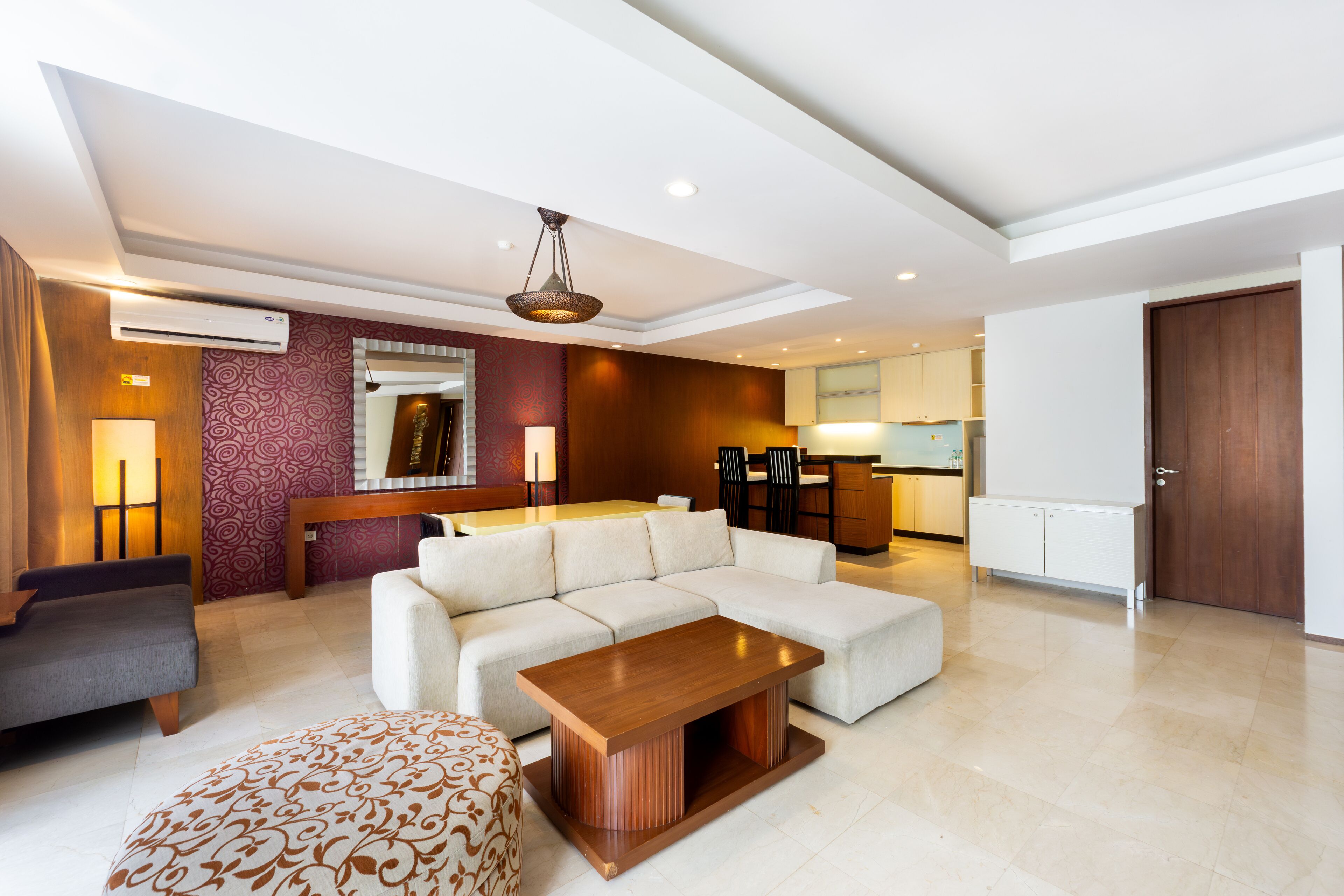 A modern living room with a sectional sofa, a coffee table, and a patterned ottoman. There is a dining area with a table and chairs, and a kitchen in the background.