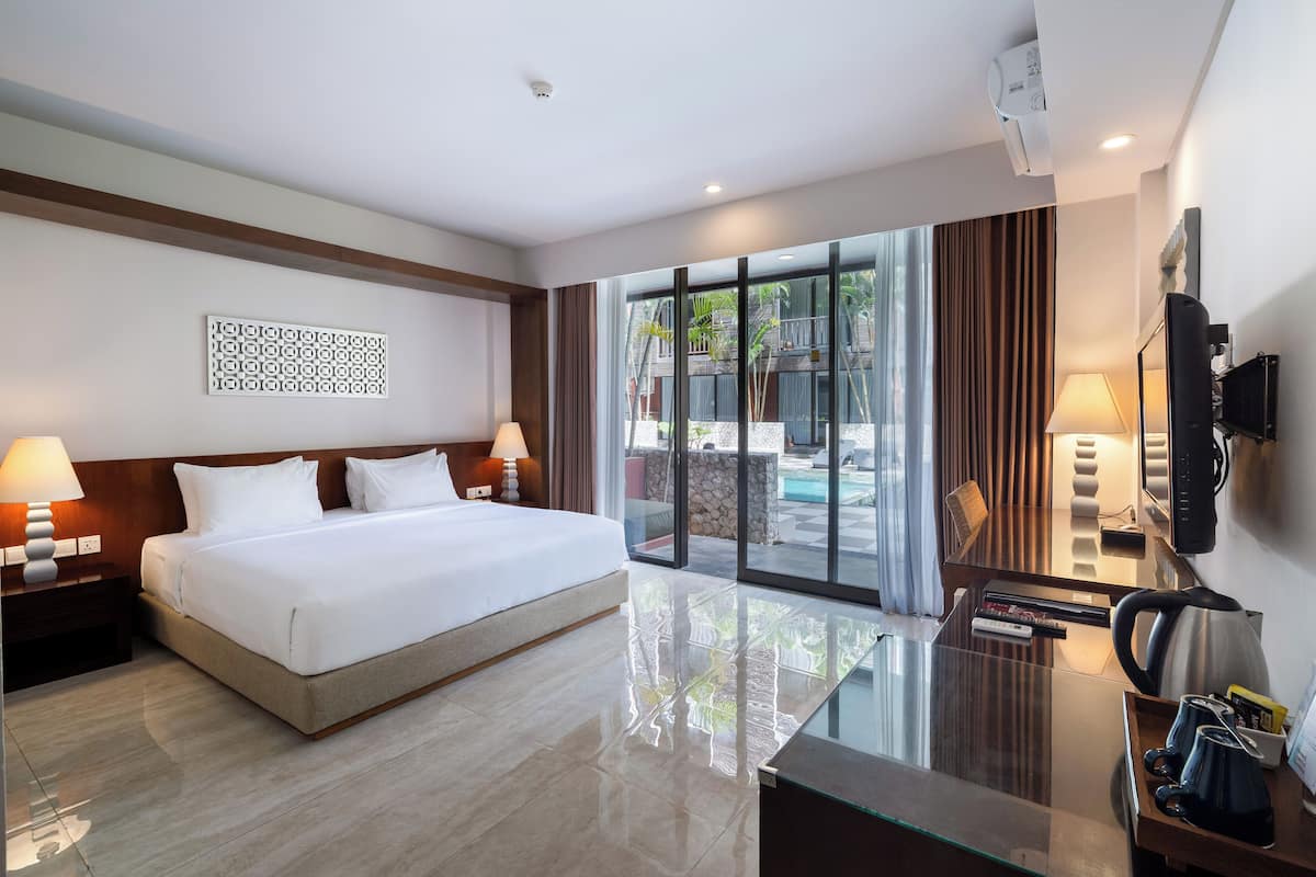 A modern hotel room with a large bed, a glass coffee table, a TV, and a view of the outdoor pool area.
