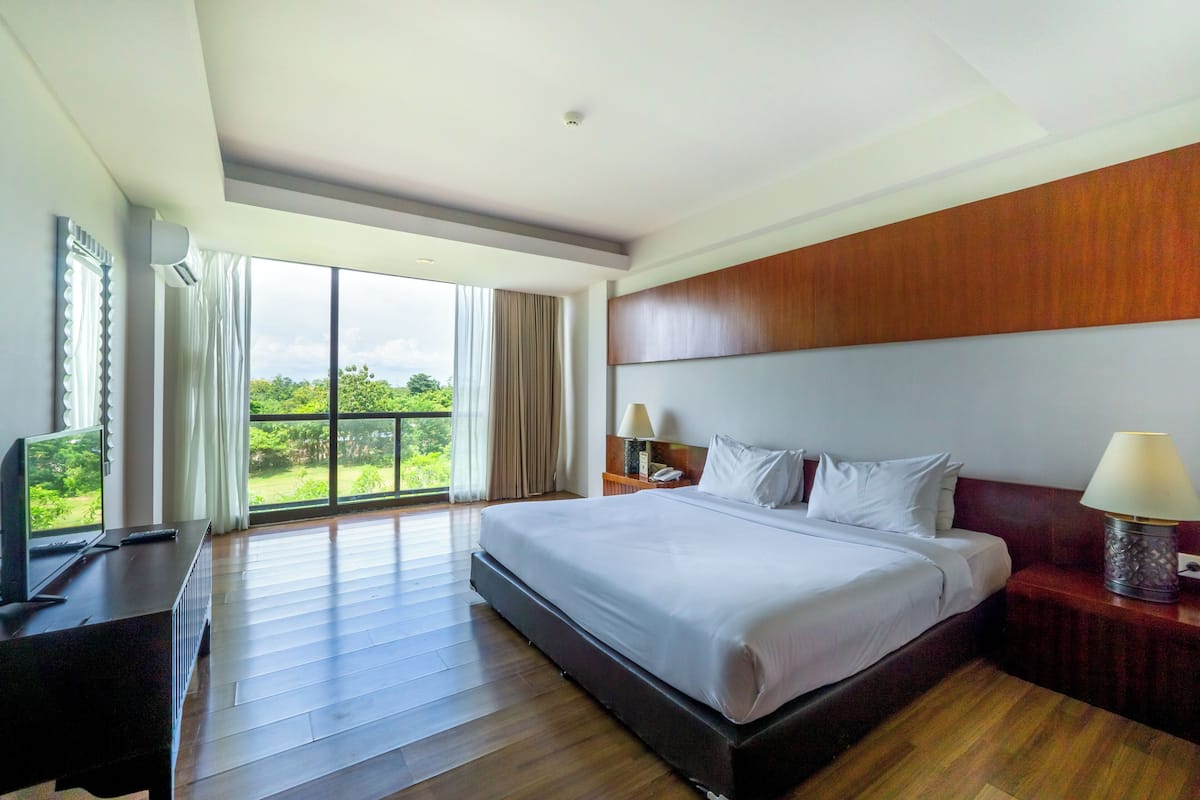A hotel room with a large bed, a desk with a computer, a nightstand with a lamp, and a view of greenery through the window.