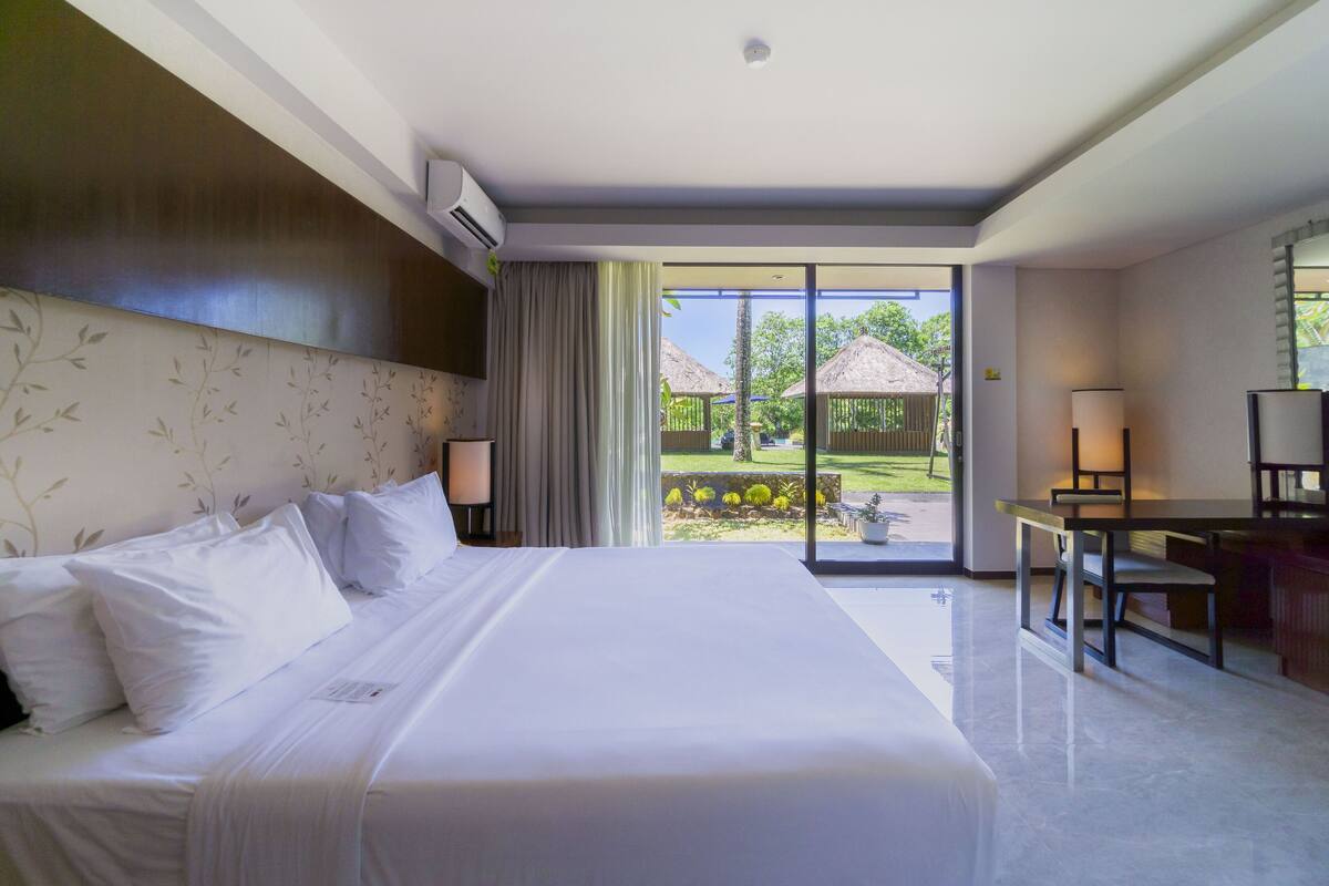 A hotel room with a large bed, a desk, and a view of a garden through a glass door.