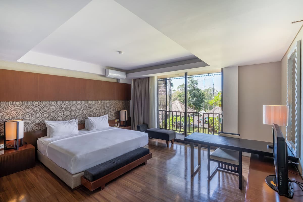 A hotel room with a large bed, a desk, a TV, and a balcony with a view of greenery.