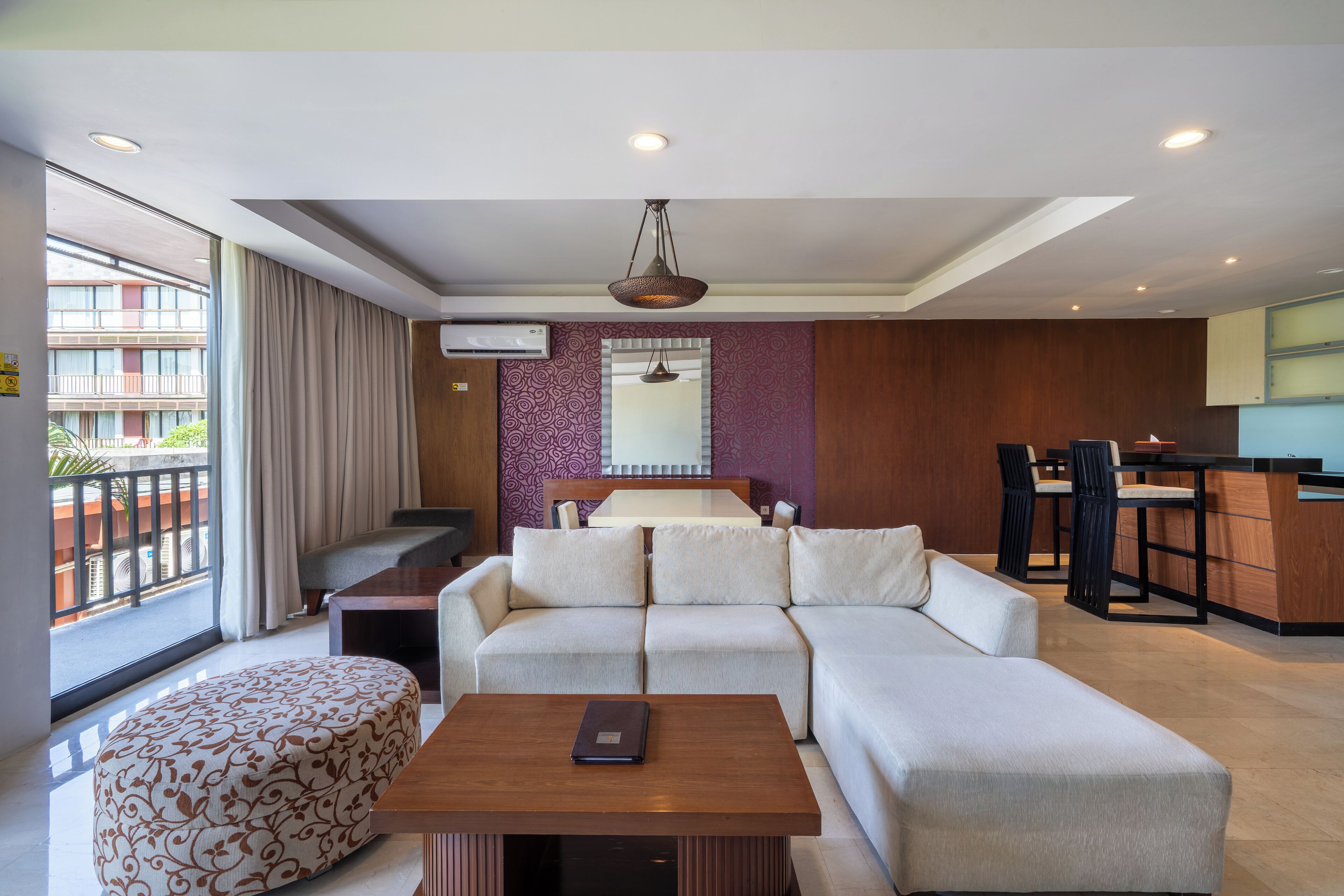 Uma sala de estar moderna com um sofá, um banquinho e uma mesa de centro de madeira. Há uma área de jantar com uma mesa e cadeiras, e uma cozinha com um balcão e cadeiras.
