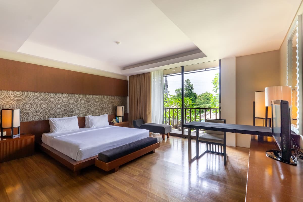 A modern hotel room with a large bed, a desk, a TV, and a balcony with a view of greenery.
