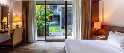 A hotel room with a bed, bedside table, lamp, and a view of a garden through a glass door.