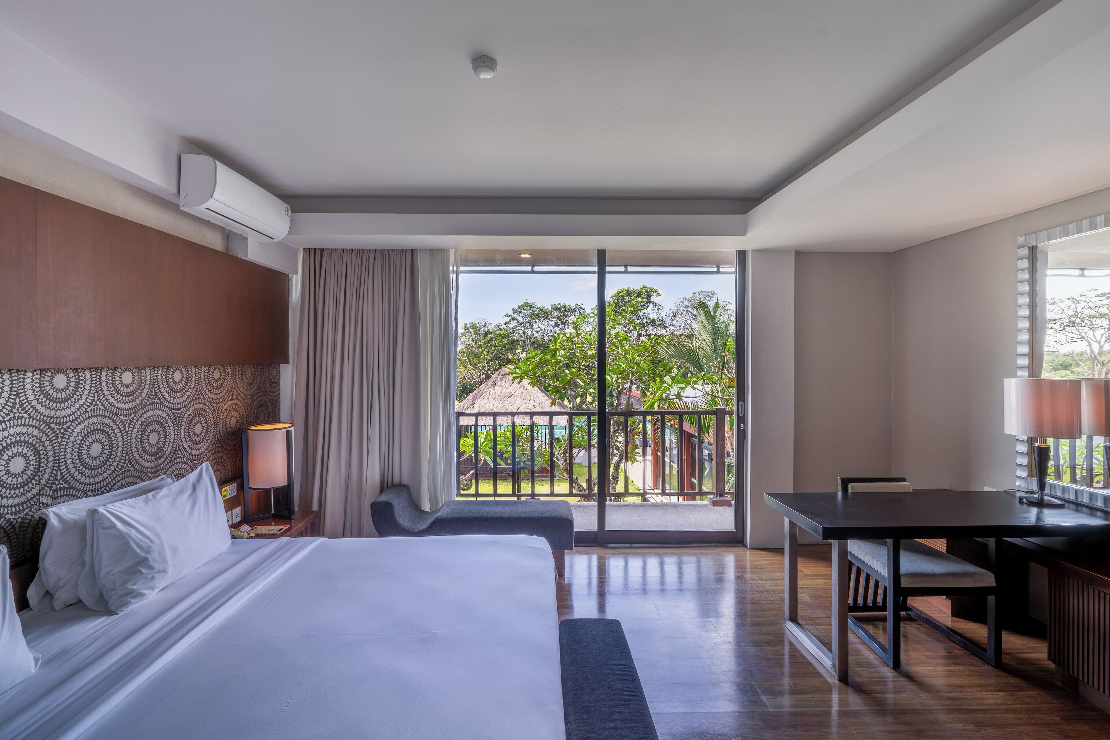 A hotel room with a bed, a desk, a chair, a lamp, and a view of a garden through a large window.