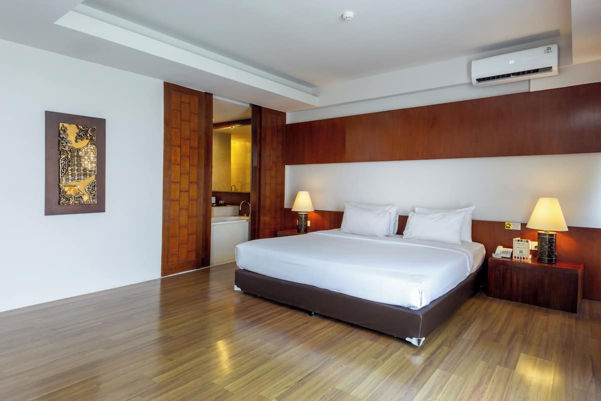 A hotel room with a large bed, wooden headboard, two bedside tables with lamps, an air conditioning unit, and a wooden floor.